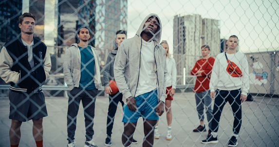 Portrait of a Handsome Athletic Black Man in a Hoodie Posing and Looking at Camera while Standing Behind a Fence on a Rooftop Urban Parking Lot. Diverse Friends Standing Behind Him