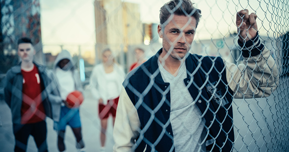 Portrait of a Handsome Caucasian Man Holding the Fence, Posing and Looking at Camera while Standing Outdoors in an Urban City Location. Stylish Diverse Friends Standing Behind Him
