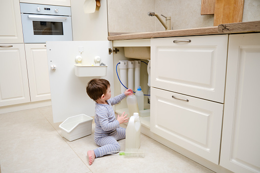 A child is playing with chemical cleaning products under the sink in the kitchen. Baby holds bottles with detergent. Kid aged about two years (one year nine months)