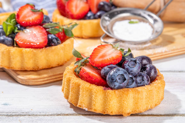 Sponge mini tart cakes with summer berry. Portioned vanilla dessert cups with fresh strawberry, blueberry, raspberry, on white wooden background copy space