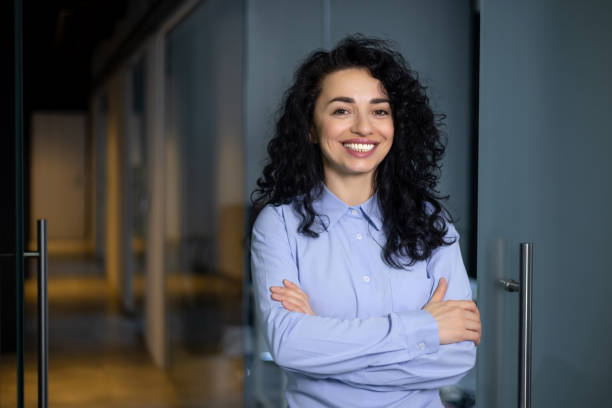 ritratto di donna d'affari felice e di successo, capo in camicia blu sorridente e guardando la telecamera all'interno dell'ufficio con le braccia incrociate, donna ispanica con capelli ricci nel corridoio - iberico sud europeo foto e immagini stock