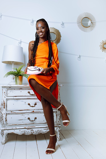 Gorgeous afro american woman in orange dress sit on vintage dresser with holiday cake in white decorated studio. Smiling birthday lady in photo zone. Birthday, holiday, celebration, festive mood.