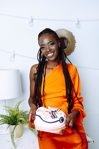 Vertical grateful, blissful, toothy smiling African American woman in orange gala dress show cake in shape of medicine box against garland in white room. Medicine doctor holiday, health day greeting