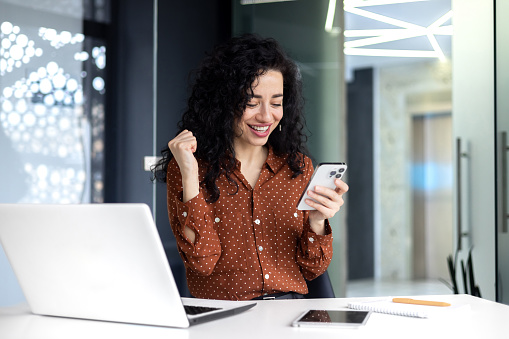 Successful satisfied business woman celebrating success and triumph, boss at workplace holding phone and hand up victory gesture, Hispanic inside office with laptop sitting at table using smartphone.