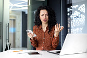 Young hispanic woman sitting in the office at the table with a laptop, using the phone and looking worriedly at the camera, raising her hands in frustration