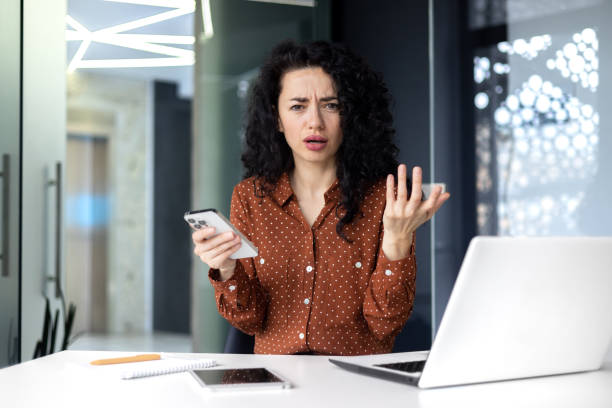 młoda latynoska kobieta siedząca w biurze przy stole z laptopem, korzystająca z telefonu i patrząca z niepokojem w kamerę, podnosząca ręce z frustracji - cell block zdjęcia i obrazy z banku zdjęć