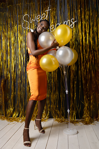 Smiling afro american woman in formal dress posing on gold decorations background in festive hall with colored balloons. Joyful birthday lady in photo zone. Birthday party, showy holiday, celebration.