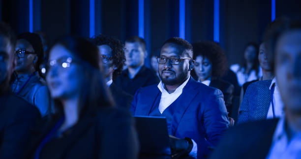 istock African Male Sitting in a Dark Crowded Auditorium at a Tech Conference. Professional Using a Laptop Computer. Specialist Watching Innovative Technology Presentation About New Software Solutions. 1482842744