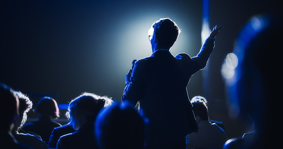 Vista trasera de un joven empresario con estilo en un auditorio oscuro y lleno de gente en una cumbre de startups. Joven hablando con un micrófono durante una sesión de preguntas y respuestas. Emprendedor feliz con el orador del evento. photo