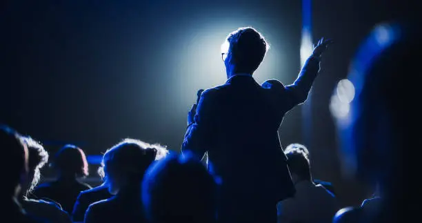 Photo of Backview of a Stylish Young Businessman in a Dark Crowded Auditorium at a Startup Summit. Young Man Talking to a Microphone During a Q and A session. Entrepreneur Happy with Event Speaker.