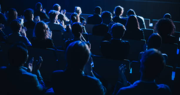 eine menge intelligenter tech-leute applaudiert im dunklen konferenzsaal während einer motivierenden keynote-präsentation. business technology summit auditorium raum voller delegierter. aufnahmen von hinten. - pressekonferenz stock-fotos und bilder