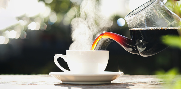 Pouring hot black coffee to white coffee cup, mug with steaming smoke of coffee on old wooden table in morning nature outdoors, garden background. Hot Drink, Beverage Concept