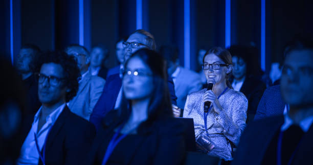 istock Female Asking a Question to a Speaker During a Q and A Session at an International Tech Conference in a Dark Crowded Auditorium. Young Specialist Expressing an Opinion During a Global Business Summit. 1482844867