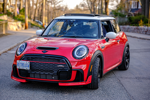 Toronto, Ontario, Canada- March 26, 2023. Chili red colour MINI COOPER on the spring sunny street  in Toronto East side, Canada. This is the third generation model F56 JCW, since BMW took over iconic brand of MINI. MINI featured in the photo is John Cooper Works model, the most powerful 2 door version. For the first time, this compact car features engine build and designed by BMW, and packs even more power and torque than previous models since 2002 to present. Original design clues and themes are still present on this brand new model. Mini has been around since 1959 and has been owned and issued by various car manufacturers.