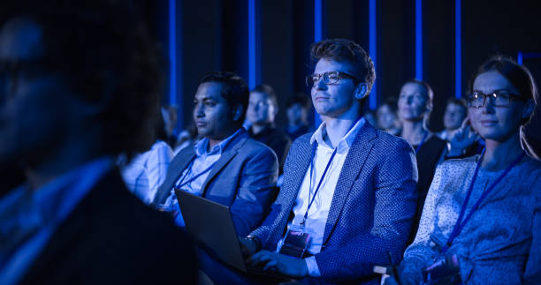 istock Male Sitting in a Dark Crowded Auditorium at a Tech Conference. Professional Using Laptop Computer. Specialist Watching Innovative Technology Presentation About New Hardware and High Tech Solutions. 1482845259