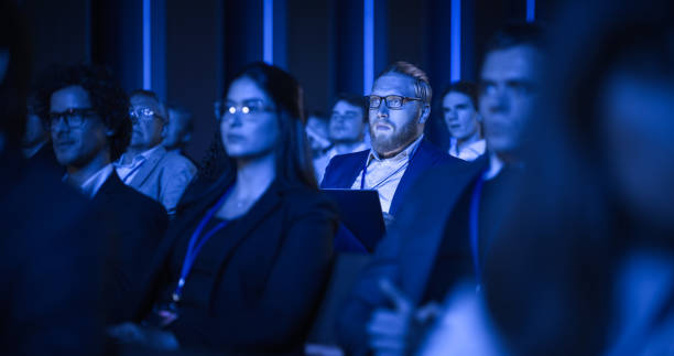 istock Male Sitting in a Dark Crowded Auditorium at an International Conference. Professional Using Laptop Computer. Innovative Presentation Concept: Technology, Science, Health, Mindfulness, Business. 1482845528