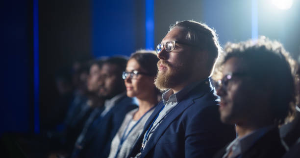 istock Young Bearded Man Attending a Political Conference, Sitting in Crowded Hall. Journalist Taking Notes and Writing an Article on Laptop Computer. Writer Listening to a Motivational Speaker. 1482845901