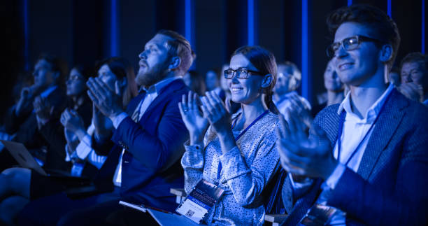 junge frau, die bei einer wissenschaftskonferenz in einem überfüllten publikum sitzt. jubeln und applaudieren die delegierten nach einer inspirierenden grundsatzrede. auditorium mit young successful specialist. - pressekonferenz stock-fotos und bilder