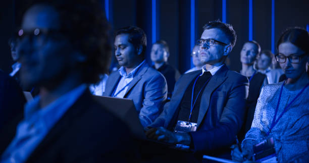istock Young Man Sitting in a Crowded Audience at a Business Conference. Corporate Delegate Watching Inspirational Entrepreneurship Presentation About Developing Markets and Financial Opportunities. 1482846539