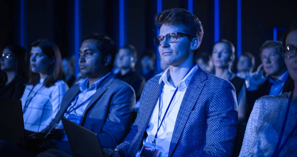 istock Young Man Sitting in a Crowded Audience at an IT Conference. Delegate Using Laptop Computer to Take Notes. Software Developer Watching Presentation About New High Tech Products and Cloud Solutions. 1482846706