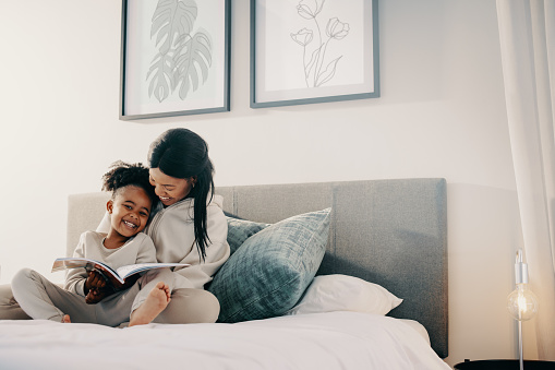 Mother and daughter spend time reading a book together, they’re smiling and sitting on a bed. Happy mom is helping her child with her reading skills. Parenting and child development.