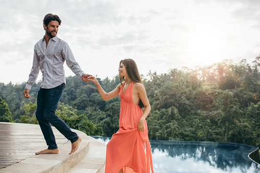 Portrait of young couple near swimming pool. Young man and woman enjoying at luxury resort poolside. Man helping woman to walking up the steps.