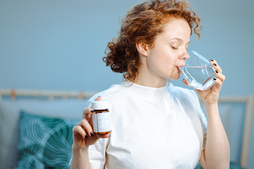Redhead woman sitting on the bed and drinking water. She is holding a bottle of medicines