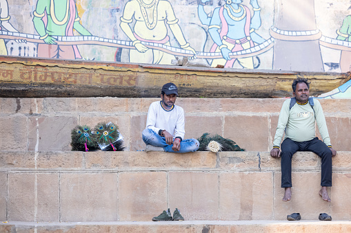Varanasi, Uttar Pradesh, India - November 2022: Portrait of Unidentified Indian male sitting on stairs with colorful painted background on ghats near river ganges in varanasi city.