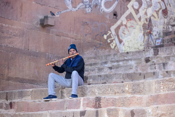 Varanasi, Uttar Pradesh, India - November 2022: Portrait of an male artist playing wooden flute on the stairs near bank of river ganges in Varanasi.