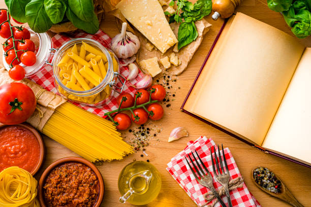 Italian pasta ingredients shot from above on wooden table. A blank cookbook is at the right of with useful copy space for text and/or logo. The composition includes raw spaghetti and rigatoni, parmesan cheese and grater, extra virgin olive oil bottle, tomatoes, salt and pepper, garlic, basil and parsley among others. High resolution 42Mp studio digital capture taken with SONY A7rII and Zeiss Batis 40mm F2.0 CF lens