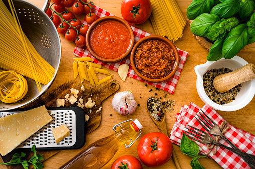 Italian pasta ingredients shot from above on wooden table. The composition includes raw spaghetti and rigatoni, parmesan cheese and grater, extra virgin olive oil bottle, tomatoes, salt and pepper, garlic, basil and parsley among others. High resolution 42Mp studio digital capture taken with SONY A7rII and Zeiss Batis 40mm F2.0 CF lens