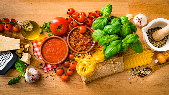 Italian pasta ingredients shot from above on wooden table. The composition includes raw spaghetti and rigatoni, parmesan cheese and grater, extra virgin olive oil bottle, tomatoes, salt and pepper, garlic, basil and parsley among others. High resolution 42Mp studio digital capture taken with SONY A7rII and Zeiss Batis 40mm F2.0 CF lens