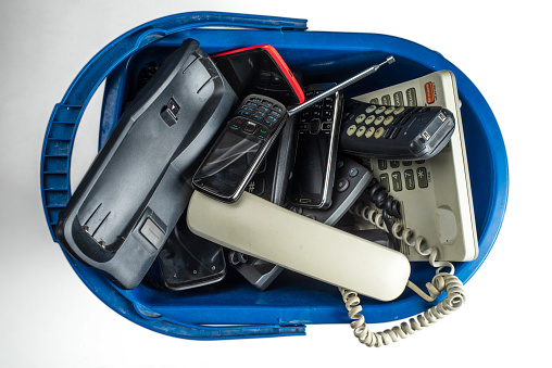 Old desk phones, cordless phones, cell phones, and smartphones in a trash can are isolated on a white background.