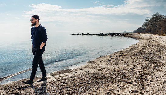 cool young man with a beard, sunglasses and a cap, walking along the shore, hands in pockets, laughing dynamically on the beach with a blue sky.