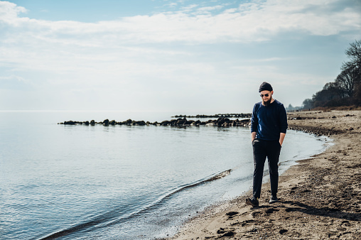 cool young man with a beard, sunglasses and a cap, walking along the shore, hands in pockets, laughing dynamically on the beach with a blue sky.