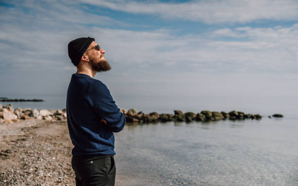 istock Cool young man with a beard, sunglasses and a cap is standing on the beach 1482859011
