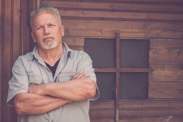 One old senior man standing outdoor against wooden tiny house in home garden looking thoughtful with crossed arms. Elderly retired lifestyle people. Handsome retired male alone outside thinking stock photo