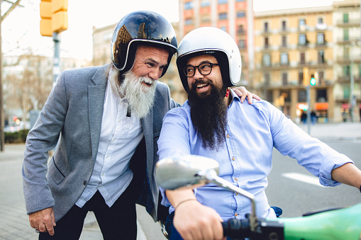 Two bearded business casual men in the street of Barcelona, next to the Tetuan square using motor scooter. In this photo they sharing moments of positive emotion and togetherness.