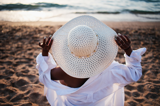 Back view african-american woman in white shirt holding her white straw hat on sandy beach looking at the horizon, no face, unrecognizable. Vacation, travel, hot season, summer vacation, relax