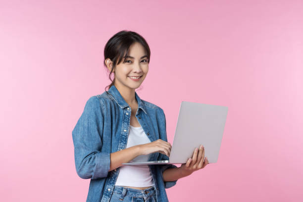 istock Young Asian college student working with a laptop computer, looking directly at the camera in a studio shot on a pink background 1482862911