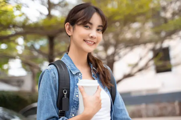 Photo of Portrait of young Asian woman student with coffee and backpack outdoor