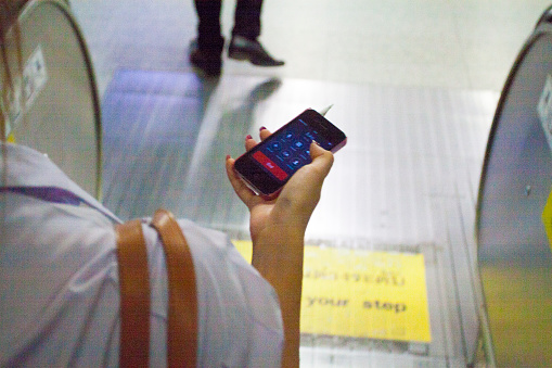 View over shoulder of young thai woman using mobile with headset and earphones for phone talk while moving down on escalator into subway station in Bangkok Ladprao. Screen is showing phone call elements
