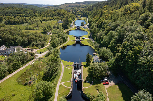 Aerial drone view of Crinan Canal, in Argyll & Bute, on the west coast of Scotland. The canal opened in 1801 and runs 14km from Crinan and Ardrishaig. The image shows the connection of canal locks, with two boats travelling through a lock to get to a different level of the canal.