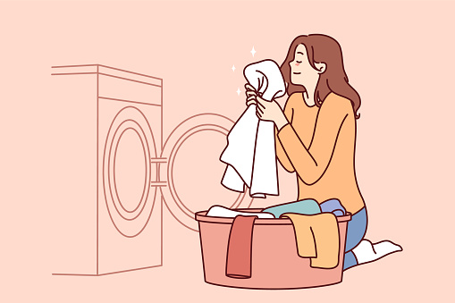 Housewife woman sits near washing machine and inhales fragrant smell freshly washed towel after using good laundry detergent. Girl takes out washed clothes from washing machine putting them in bowl