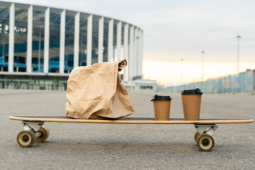 Takeaway food in paper bag and two paper cups with hot drink placed on longboard against blurred cityscape background