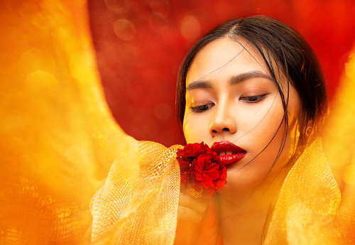 Asian Beauty Gil Portrait with Red Full Lips smelling Flower. Chinese Woman with Smooth Skin Makeup. Beautiful Japanese Model with golden Fabrics over art background