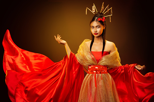 Asian Empress in Red Silk Dress with Golden Jewelry. Beautiful Chinese Girl in Traditional Wedding Costume Clothes. Japanese Model in Fantasy Bride Gown over Dark Studio Background