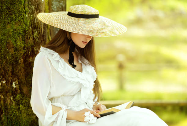 Romantic Girl in White Dress and Straw Sun Hat reading Book sitting under Tree. Young Woman studying in Spring Park. Old Fashioned Elegant Lady outside stock photo