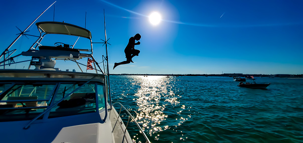 Young boy jumping into the crystal clear turquoise water from the tower of a boat on the first weekend of summer