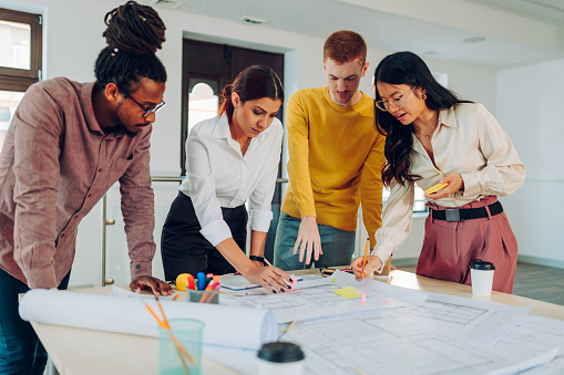 Multiracial engineer team working at the office table with construction plans and architect equipment while sketching and planning construction project. Focus on an asian female team leader.